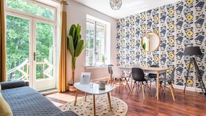 A room with a large window, a dining table with chairs, a round mirror, and a potted plant.