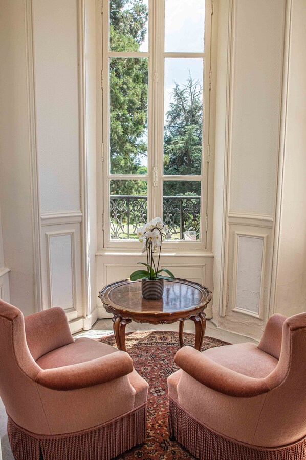 A room with two pink armchairs, a round table with a vase of flowers, and a large window with a view of trees.