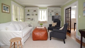 Een woonkamer met een witte bank, een grijze fauteuil, een bruin poef en een televisie op een witte kast.