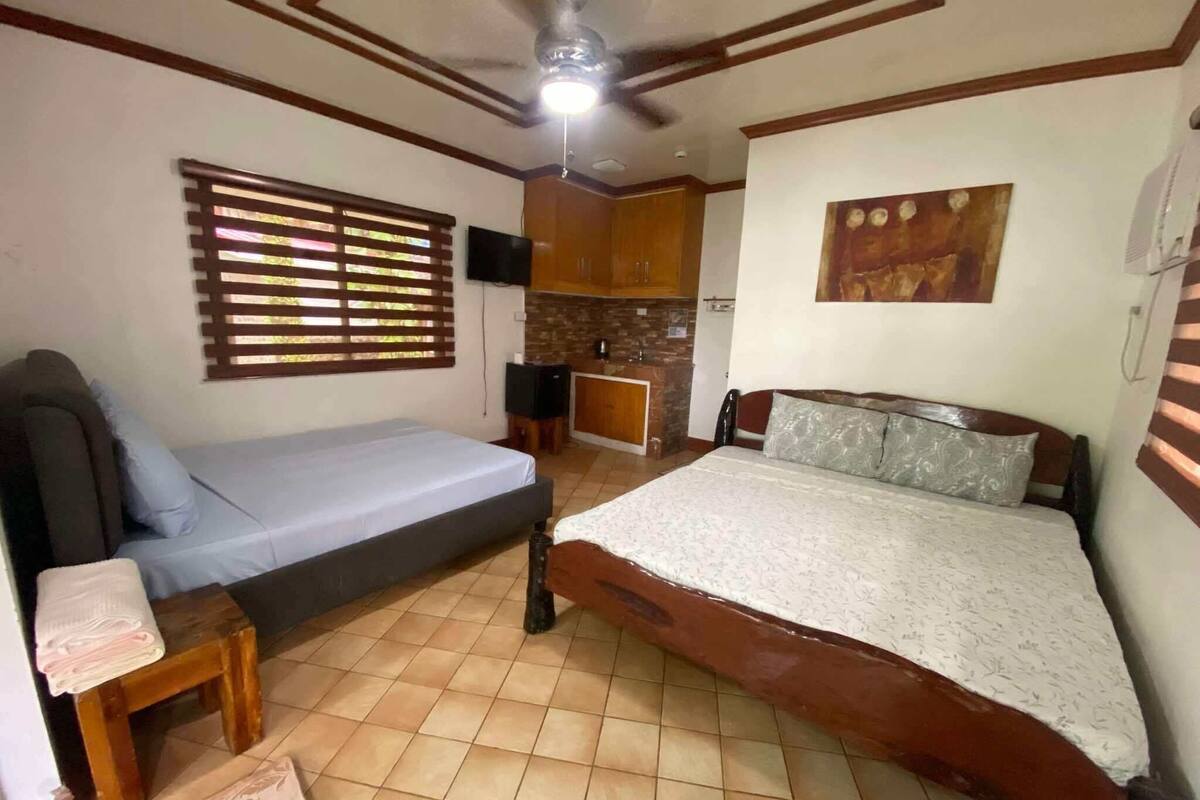 A bedroom with a bed, a sofa, a small table, a TV, and a kitchen area.