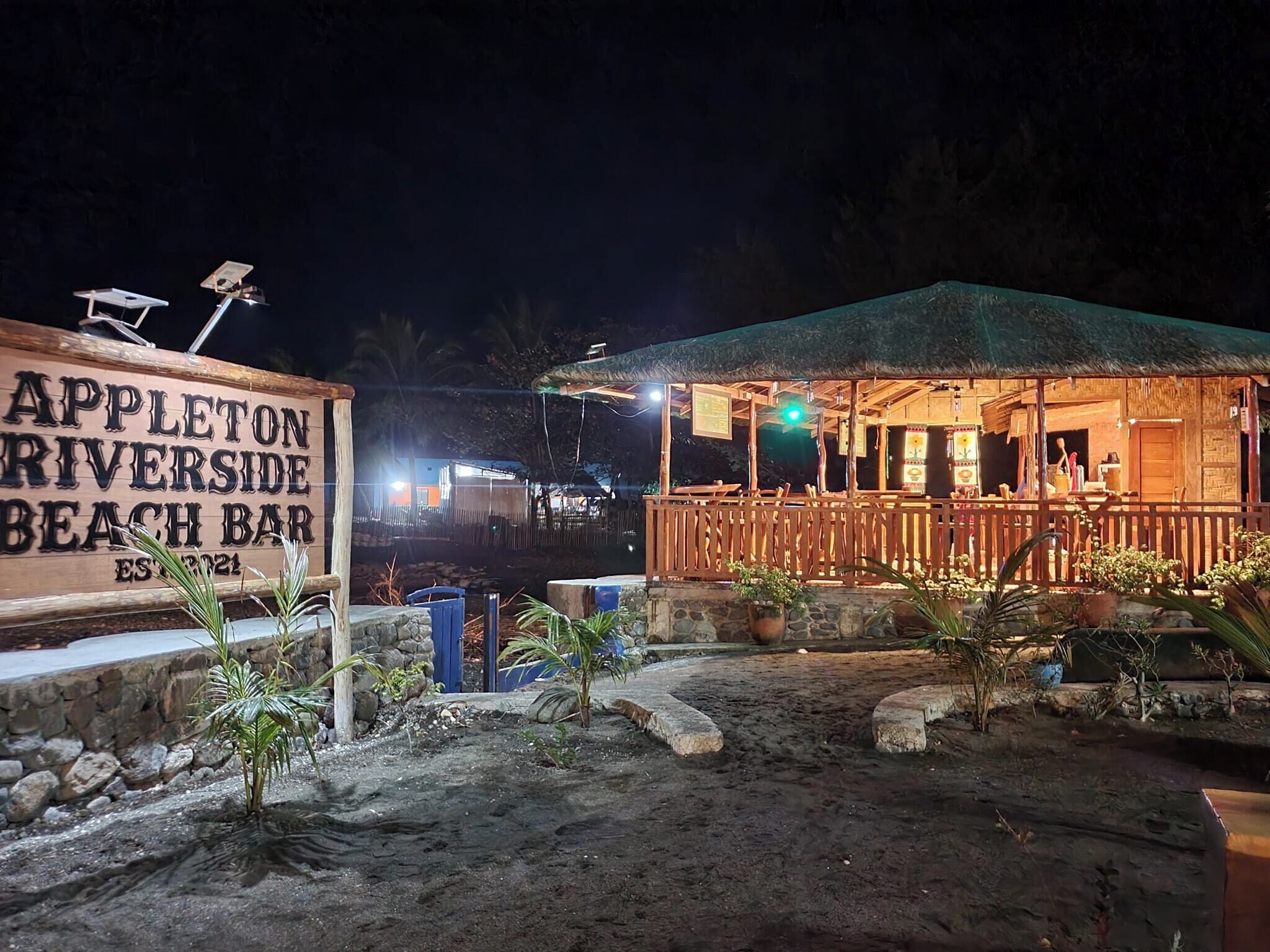 A beach bar with a wooden signboard, outdoor seating, and a thatched roof structure.