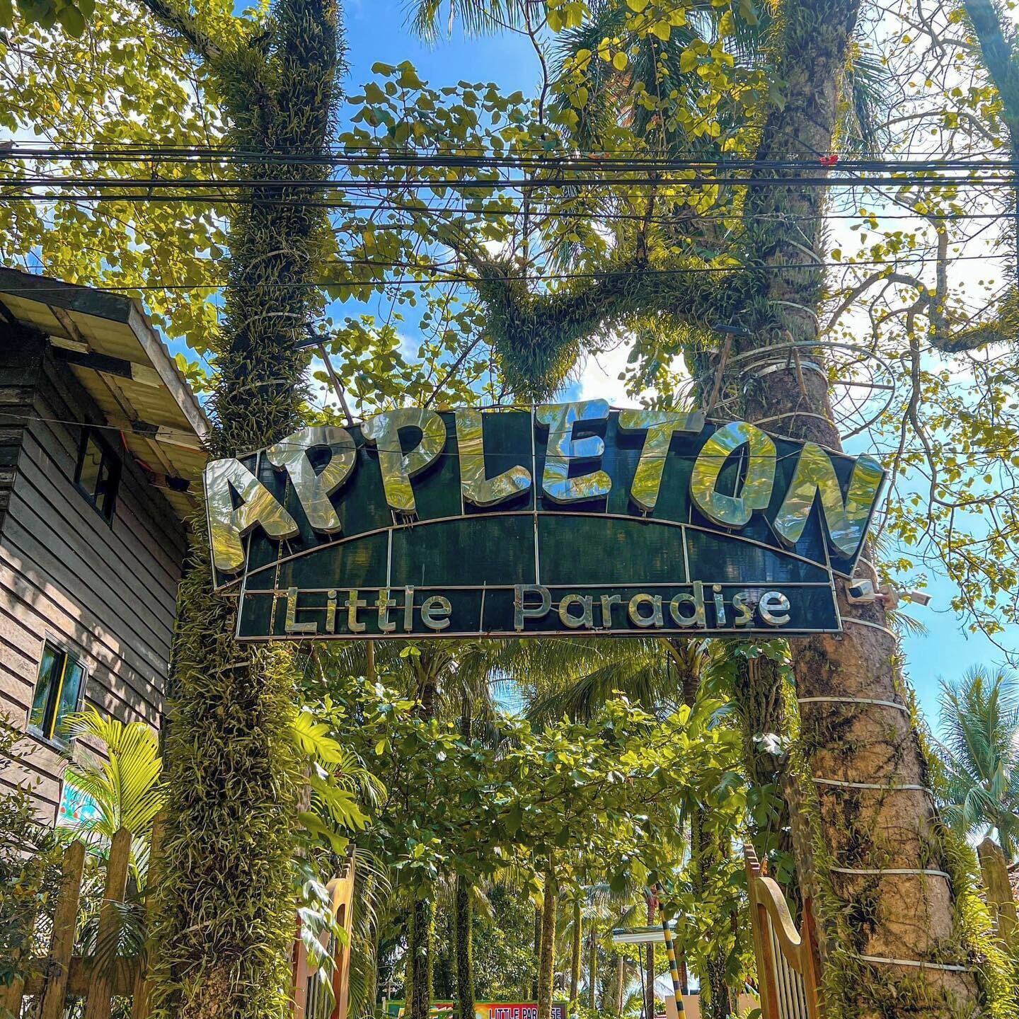 Ein Schild mit der Aufschrift 'Appleton Little Paradise', im Hintergrund ein rustikales Holzhaus.