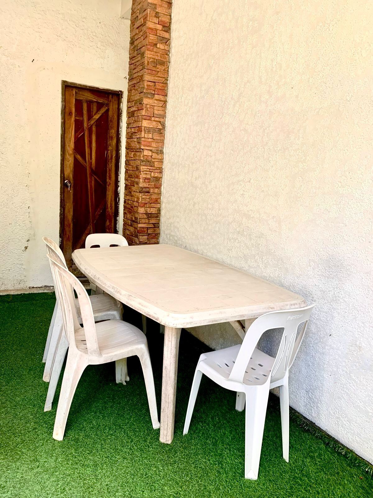 Una mesa rectangular al aire libre con cuatro sillas blancas de plástico, frente a una pared blanca con una puerta de madera y un pilar de ladrillo.