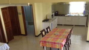 A dining area with a table covered in a checkered tablecloth, surrounded by chairs. There is a kitchen area with cabinets and appliances in the background.