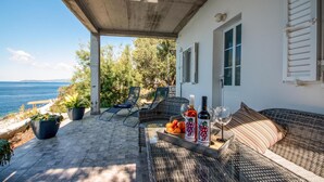 A covered outdoor seating area with wicker furniture, a table with a bottle of wine, glasses, and a bowl of fruit, overlooking a body of water. - Cozy home in Potomje with WiFi (Potomje)