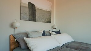 A neatly made bed with a grey and white color scheme, a wooden headboard, and a wall-mounted painting of a natural scene. - Newly renovated apartment, lake view, directly on the south beach, WLAN & beach chair included (Burg auf Fehmarn)