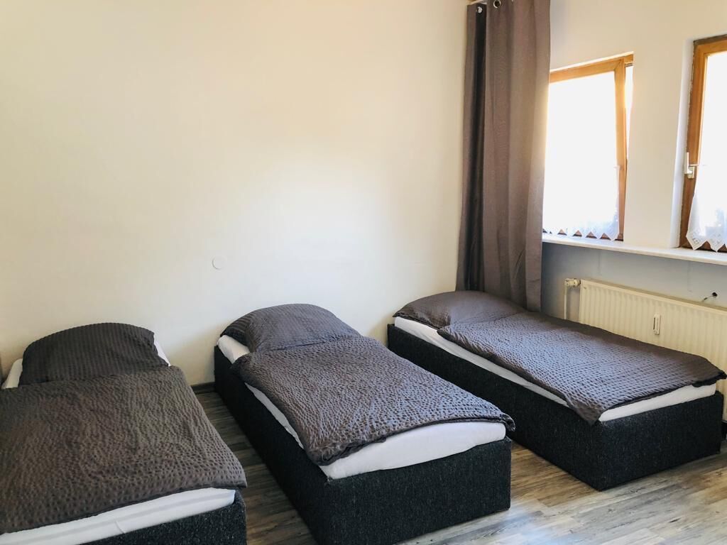 Ein Zimmer mit drei an einer Wand stehenden Einzelbetten, einem Fenster mit Vorhängen und einem Heizkörper.