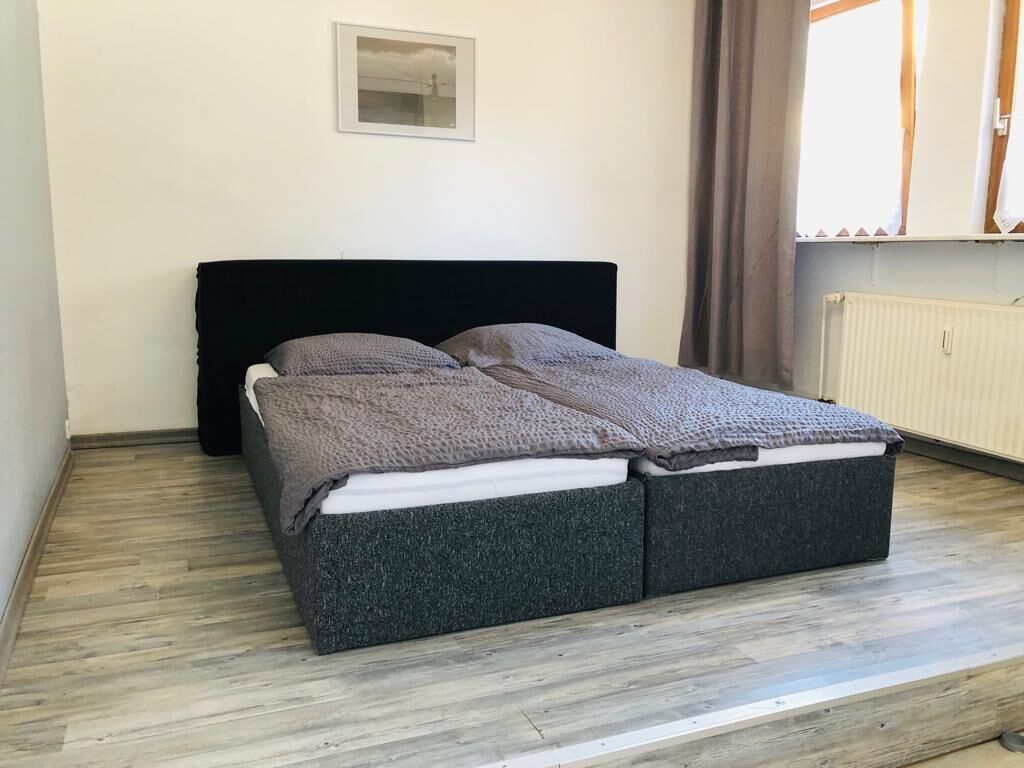 Una cama con cabecera negra, ropa de cama gris y un caja gris debajo del colchón.