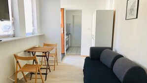 A compact living space with a dining area featuring wooden chairs and a table, a black sofa, and a kitchen visible in the background.