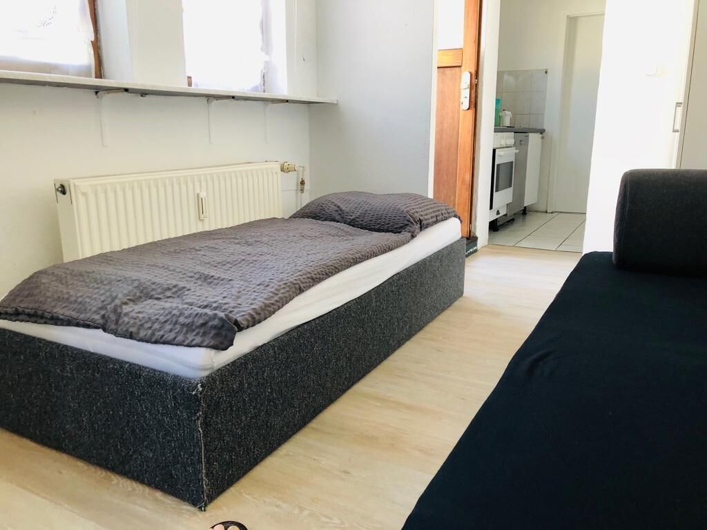 A room with a bed, a black sofa, a radiator, and a kitchen area in the background.