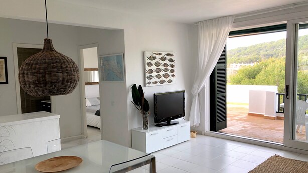 Ein modernes Wohnzimmer mit einem Glastisch, einem Fernseher auf einem weißen Schrank und einem Balkon mit Blick auf Grünflächen.