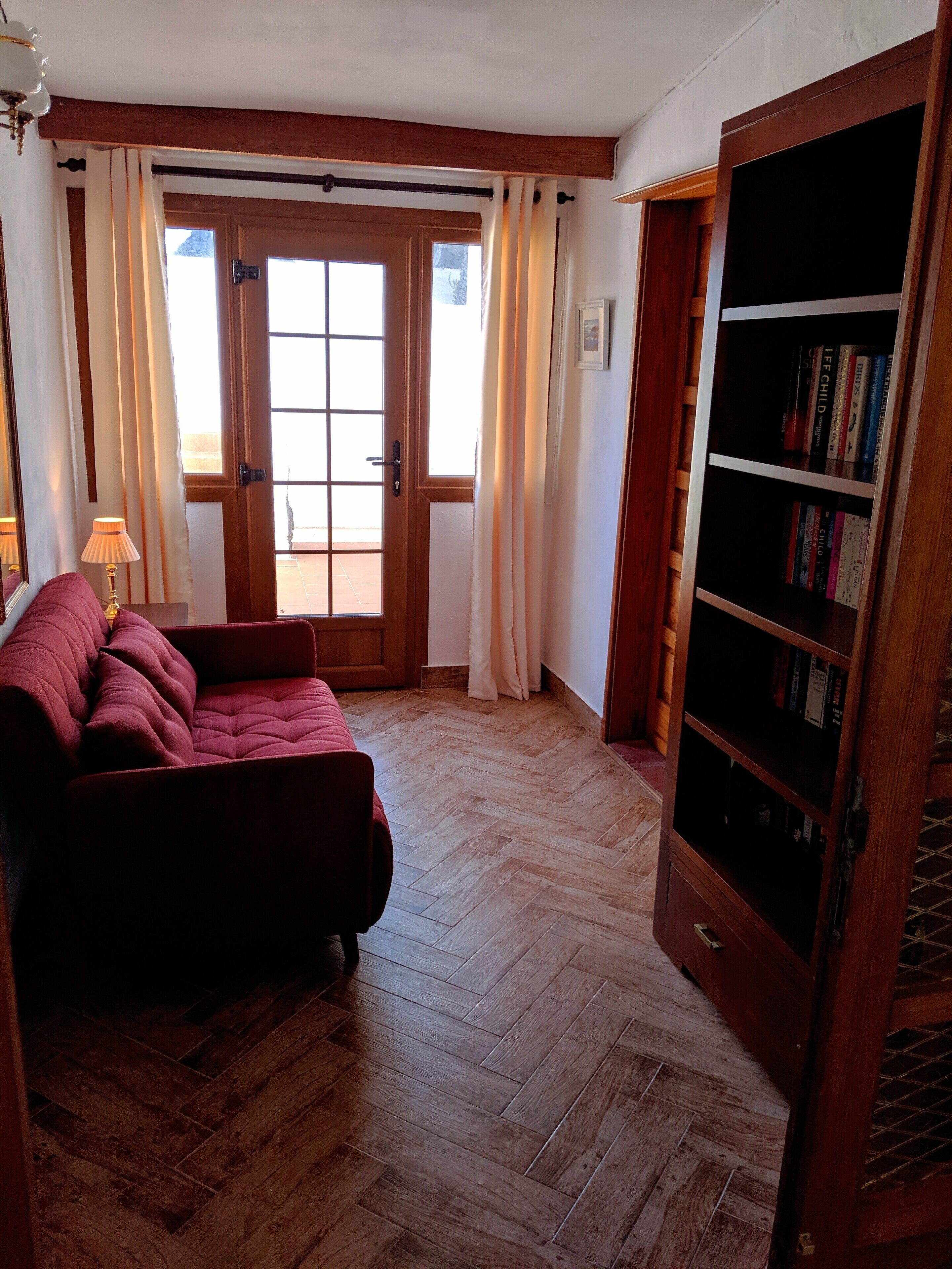 A room with a herringbone wooden floor, a red sofa, a wooden bookshelf, and a door leading outside.