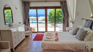 Quarto com cama, cômoda, cadeira e vista para o oceano através de grandes janelas.