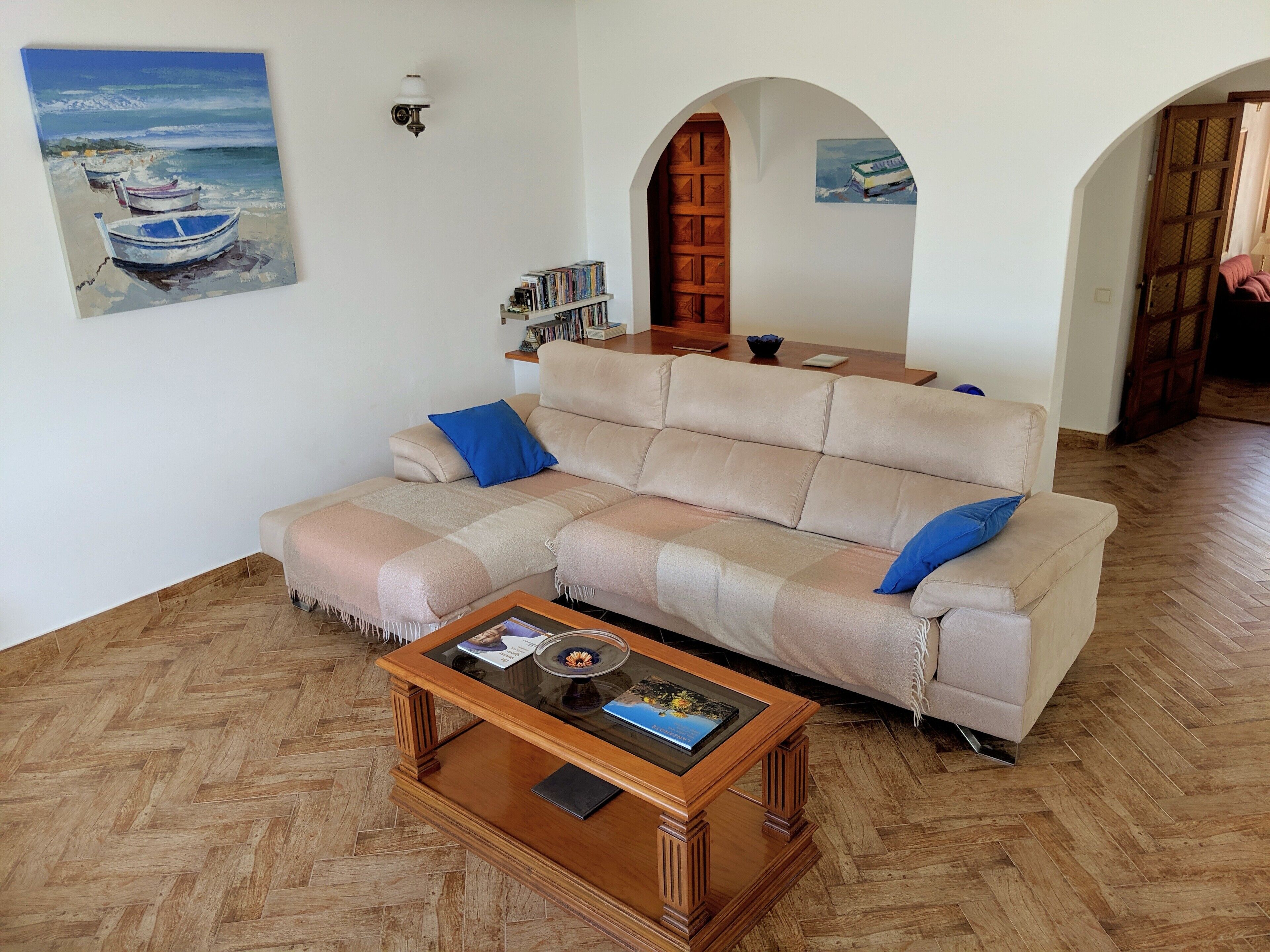 A living room with a beige sofa, a wooden coffee table, a bookshelf, and a painting of boats on the wall.