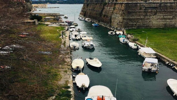 A marina with boats docked along a stone wall, a grassy area, and a distant building. - Pelo Appartment corfu (Corfu)