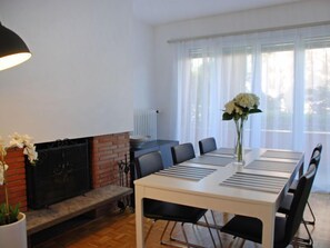 Een eetgedeelte met een witte tafel, zwarte stoelen, een open haard en een raam met dunne gordijnen.