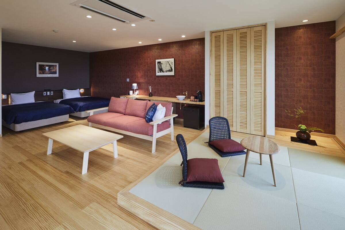 A modern hotel room with a sofa, two chairs, a small table, a desk with a lamp, and a large window with blinds.