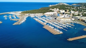 Une vue aérienne d’une marina où sont amarrés de nombreux bateaux, avec un grand ensemble d’immeubles et un paysage vallonné en arrière-plan.
