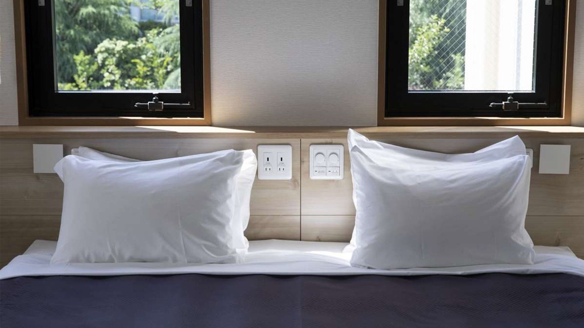 Un lit avec des oreillers blancs et une tête de lit en bois, deux prises électriques et une fenêtre donnant sur un paysage verdoyant.