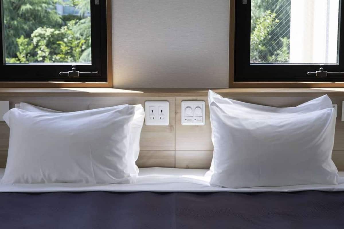 A bed with white pillows and a wooden headboard, two electrical outlets, and a window with a view of greenery.