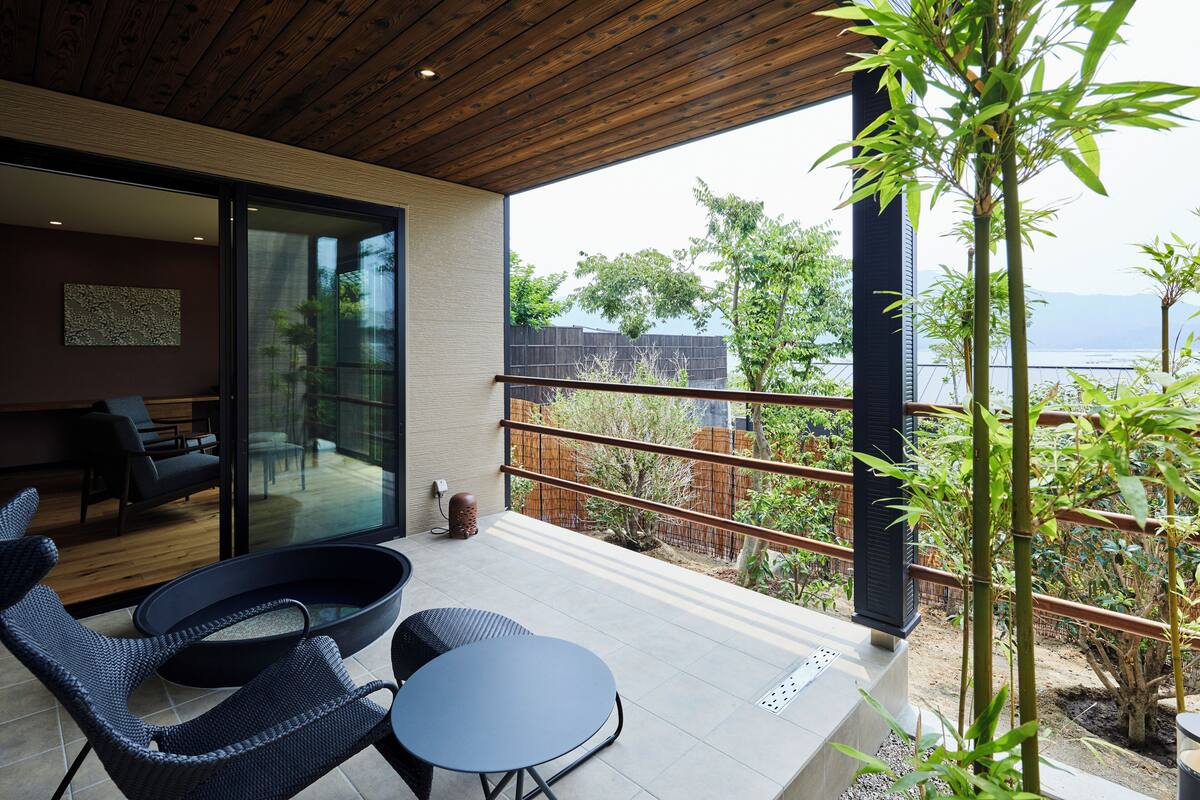 Een modern balkon met rieten meubilair, een glazen deur naar een zithoek, en uitzicht op groen en een verafgelegen landschap.