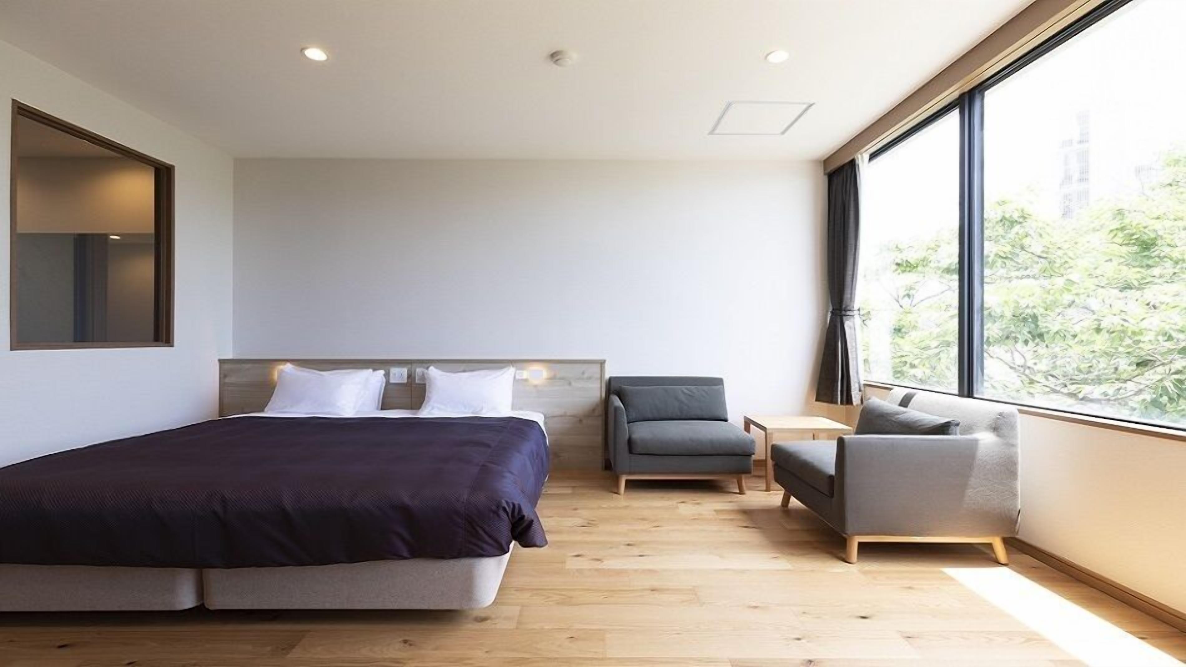 Une chambre d’hôtel moderne, dotée d’un grand lit, d’un petit canapé, d’un fauteuil et d’une grande fenêtre donnant sur l’extérieur.