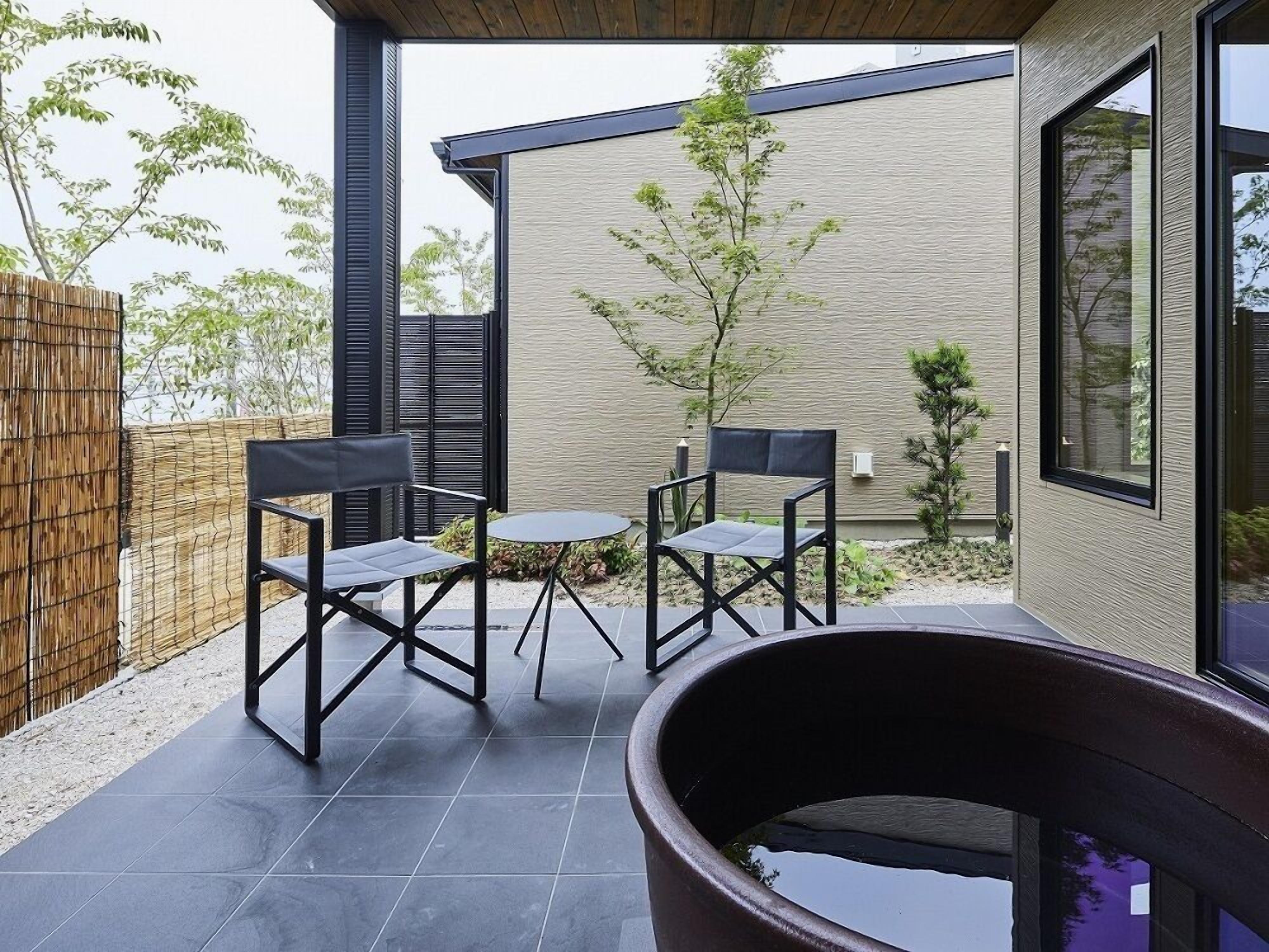 Un patio extérieur moderne avec deux chaises et une petite table, un grand bac à plantes et un bâtiment doté d’une façade texturée et de grandes fenêtres.