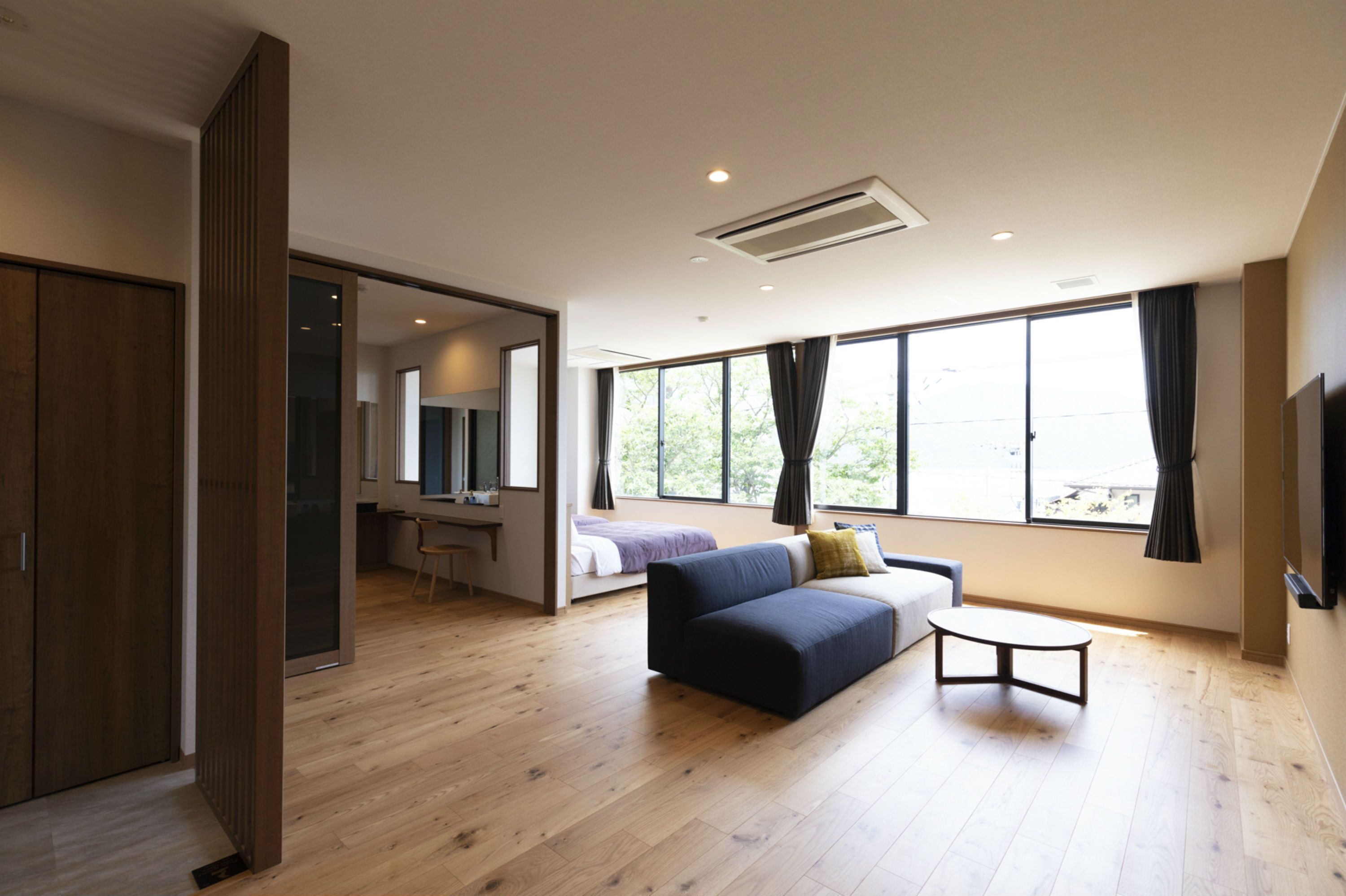 Una habitación de hotel moderna con suelo de madera, un sofá, una mesita, un escritorio, una cama y ventanas amplias con cortinas.