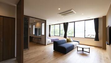 Una habitación de hotel moderna con suelo de madera, un sofá, una mesita, un escritorio, una cama y ventanas amplias con cortinas.