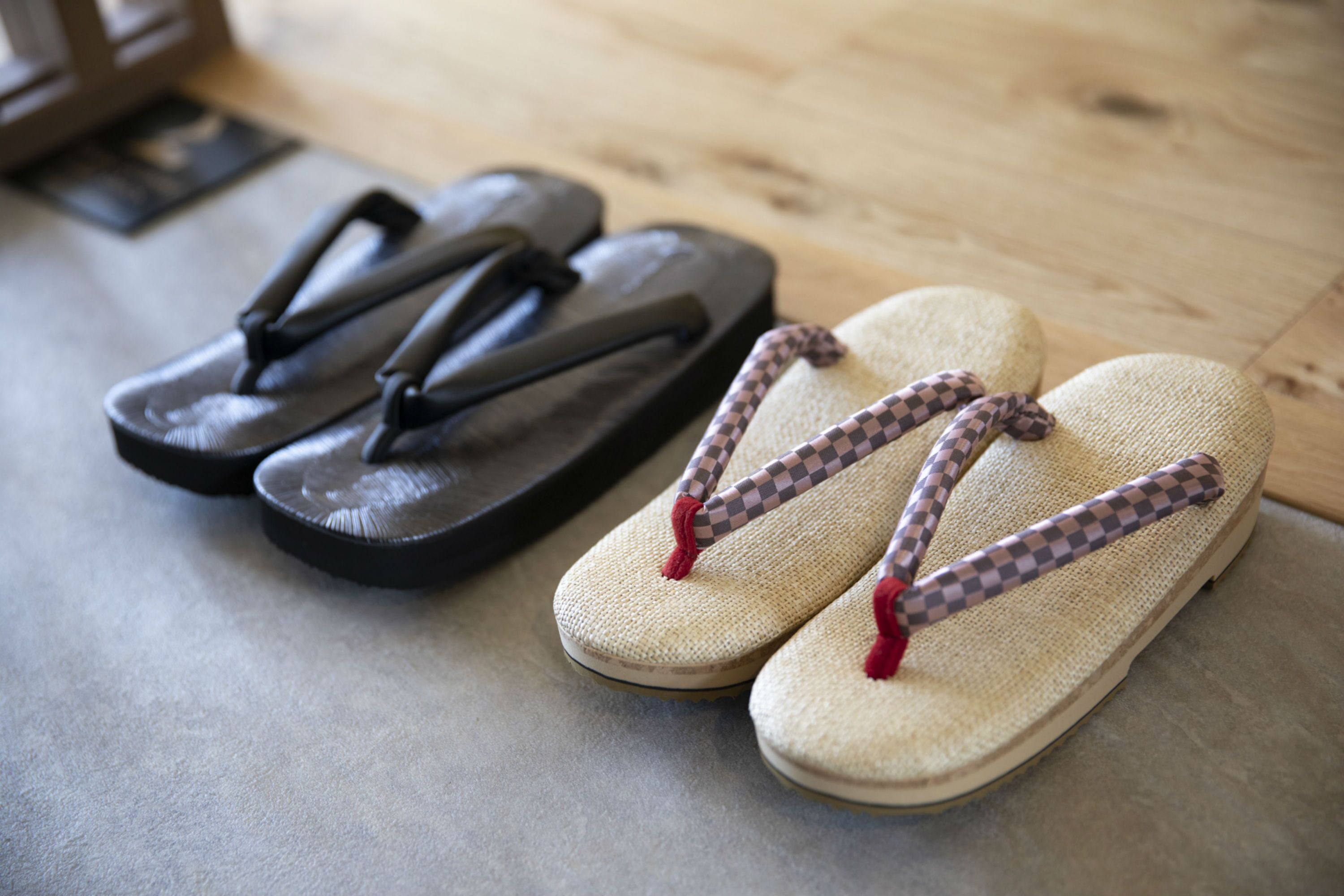 A pair of black flip-flops and a pair of straw sandals with checkered straps on a wooden floor.