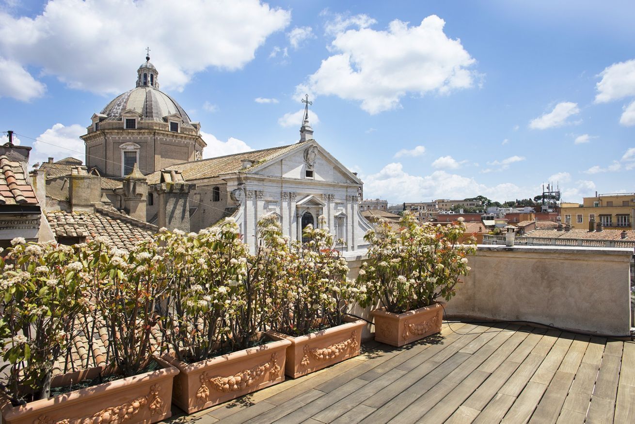 A rooftop terrace with potted plants, a building with a dome and a bell tower, and a clear blue sky.