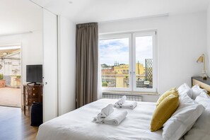 A hotel room with a bed, a TV, a window with a view of buildings, and a door leading to a balcony.