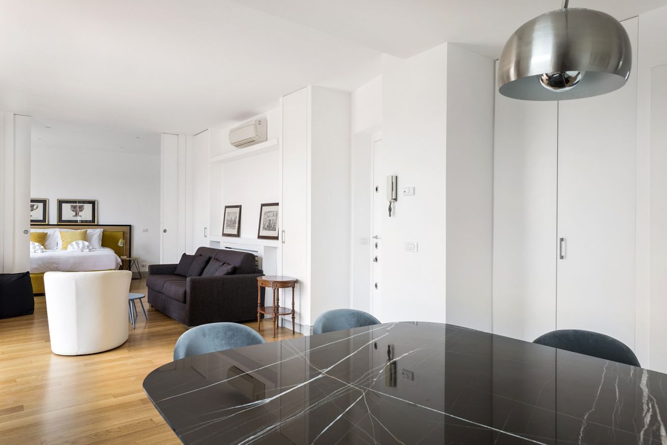 Un salon moderne avec une table en marbre foncé, un canapé et un pouf rond blanc. On y trouve également un coin repas avec une grande table et des chaises, ainsi qu’un luminaire suspendu au plafond.