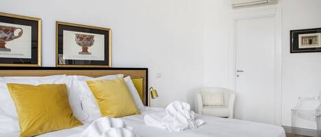 A hotel room with a bed, white bedding, yellow pillows, a white chair, and two framed pictures on the wall.