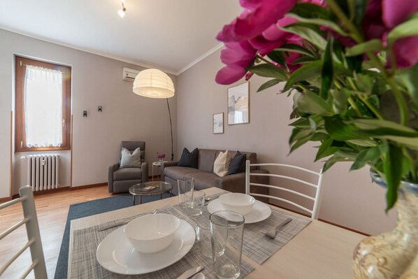 A dining area with a table set for two, a sofa, a radiator, and a window with curtains.