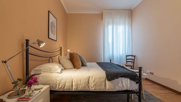 A bedroom with a bed, bedside table, lamp, chair, and a window with curtains. - iFlat | Augustus Suite near Saint Peter (Roma)