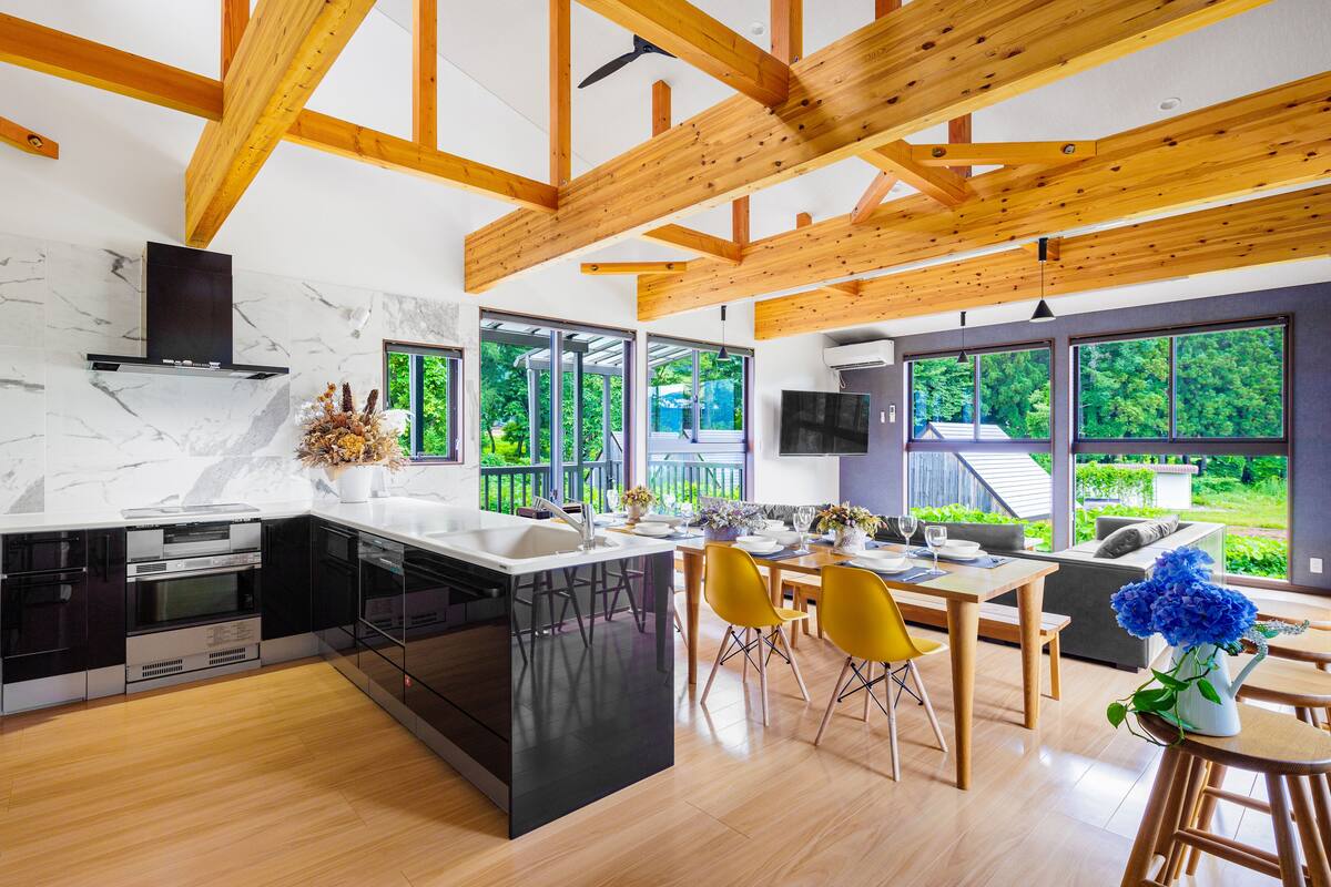 A modern kitchen with a central island, wooden beams, and large windows offering a view of greenery.
