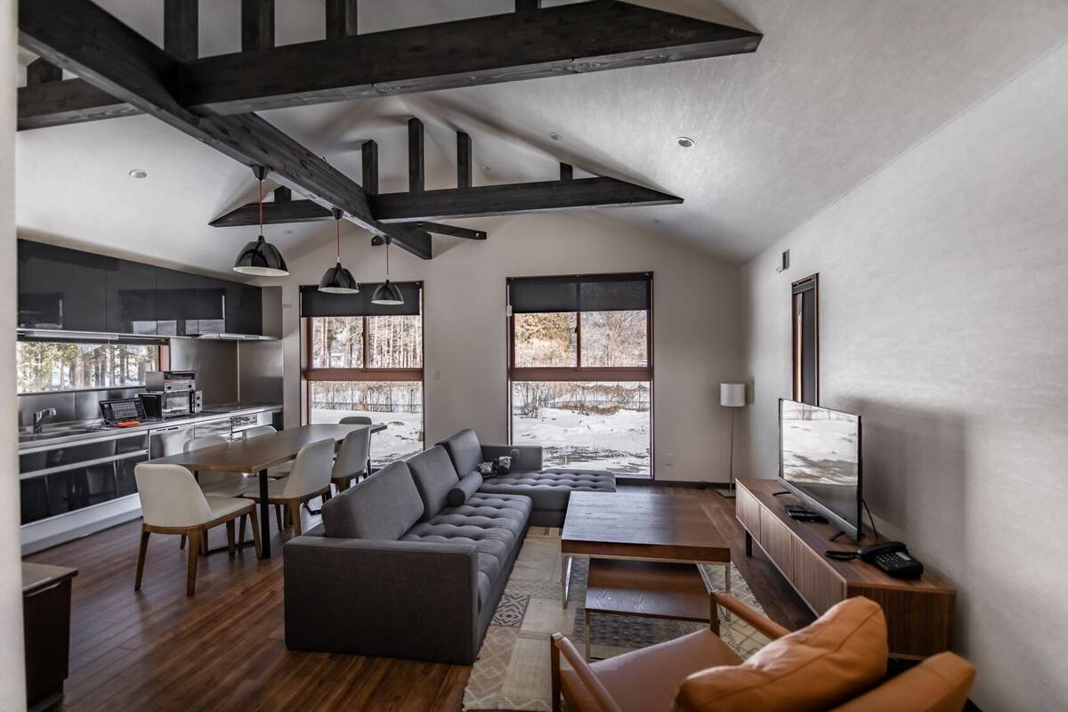 A modern interior with a kitchen, dining area, and living room, featuring exposed beams, large windows, and a flat-screen TV.