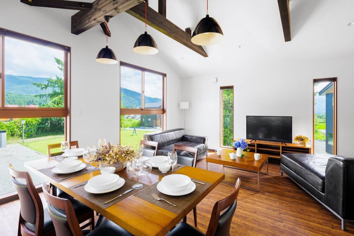 A dining area with a table set for four, a sofa, a TV, and large windows with a view of greenery.