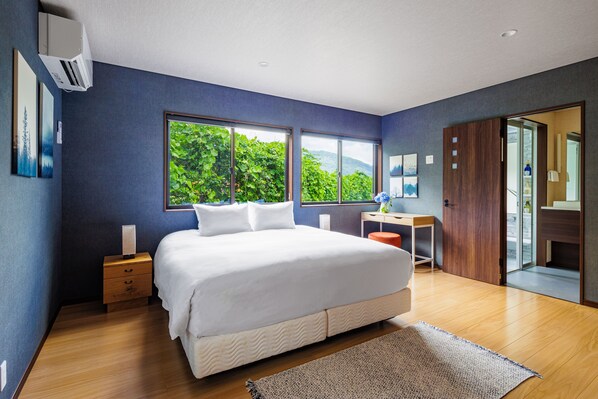 Un dormitorio con una cama grande, un escritorio y vistas a la vegetación a través de las ventanas.