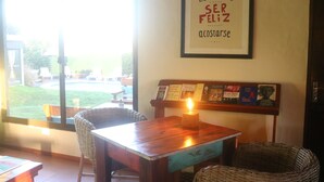 Un café acogedor con sillas de mimbre, una mesa de madera con una vela, y una pared con libros enmarcados y una cita motivacional.