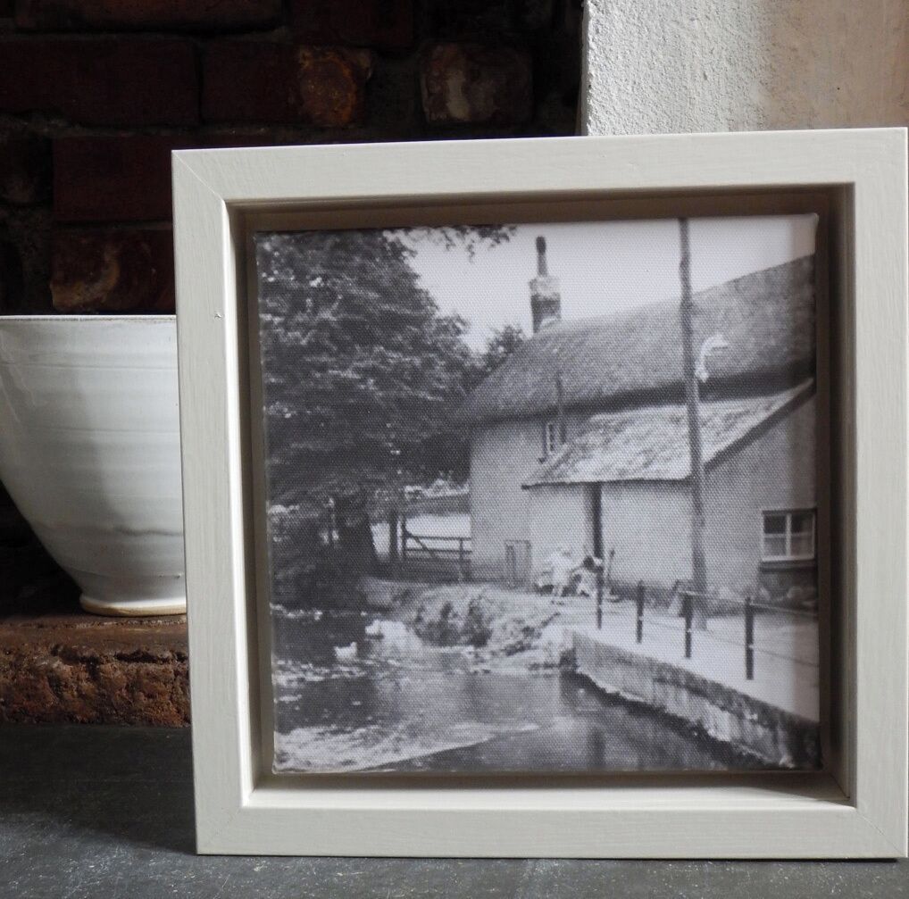 A framed black and white photograph of a house with a thatched roof, a small pond, and a wooden fence.
