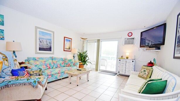A living room with a sofa, a coffee table, and a TV. There is a window with curtains and a door leading to a balcony.