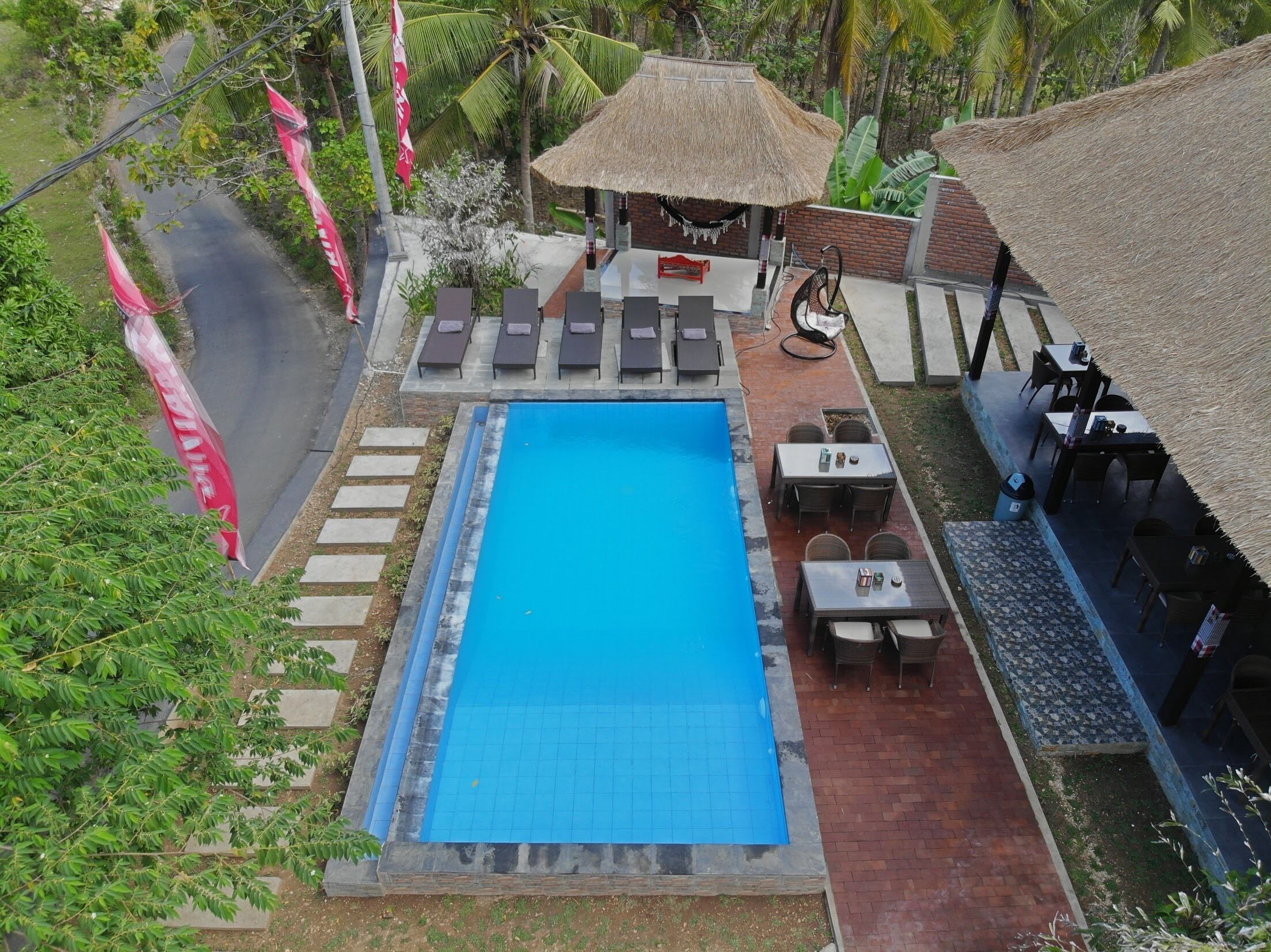 Una piscina al aire libre con sillas de descanso y mesas, rodeada de vegetación tropical y construcciones con techo de paja.
