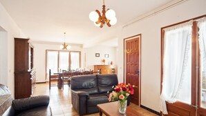 A living room with a sofa, a wooden coffee table with a vase of flowers, a dining area with a table and chairs, and a wooden door with curtains. - Villa with swimming-pool and garden.HUTG-015589 (l'Escala)