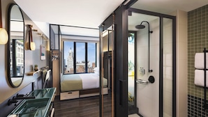 Habitación de hotel moderna con una cama grande, cabina de ducha de vidrio, lavamanos verde y vista a la ciudad.