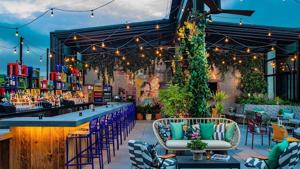 Un área de bar al aire libre con mostrador de madera, taburetes azules y una zona de estar con muebles de mimbre y cojines.