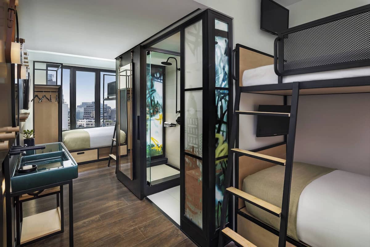 A modern hotel room with a bunk bed, a desk with a chair, a large window with a city view, and a sliding door leading to a bathroom.