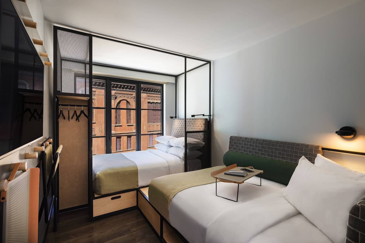 A modern hotel room with two beds, a wardrobe, a desk, and a view of a brick building.