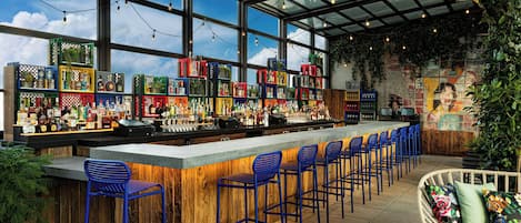 Uma área de bar com um balcão comprido, banquetas azuis e uma variedade de garrafas e copos.